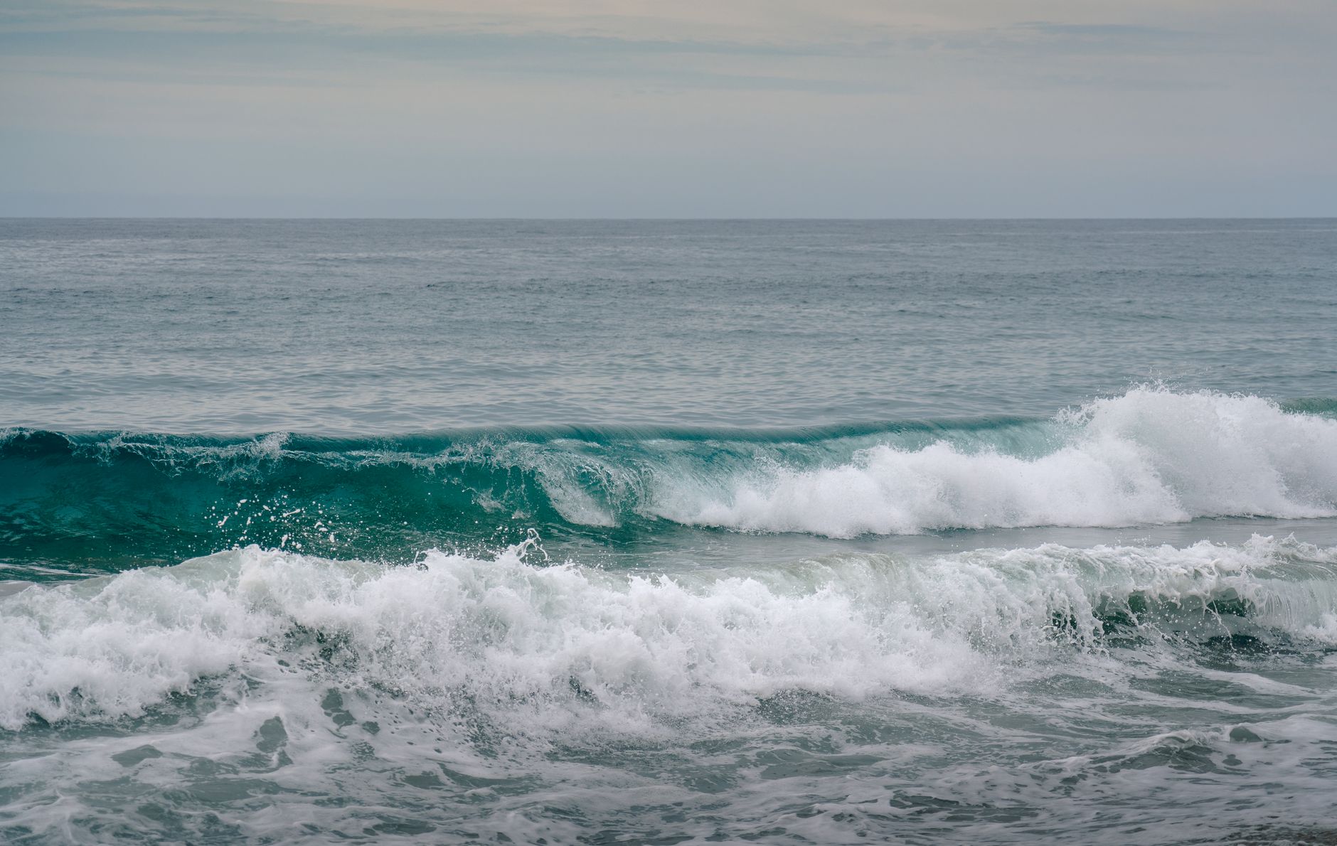 Tranquil view of ocean waves crashing onto the sandy shore under a calm cloudy sky.
